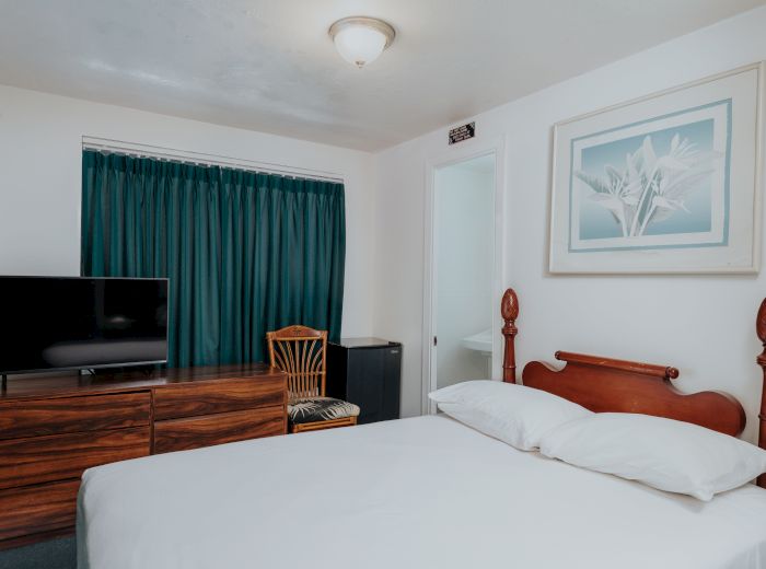 A simple bedroom with a bed, teal curtains, a wooden dresser with a TV, a chair, and a framed picture on the wall ends the sentence.