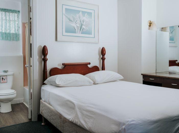 A small bedroom with a bed, pillows, framed artwork, a dresser with a mirror, and an open door revealing a bathroom with a toilet.