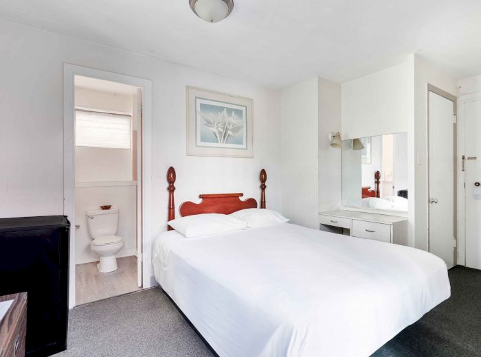A bedroom with a bed, white linens, a bedside table, a painting, and an adjacent bathroom with a visible toilet and window.