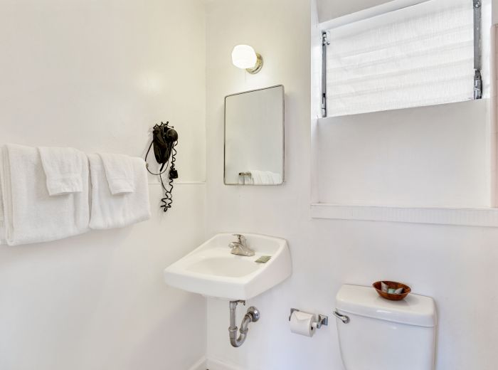 A clean white bathroom with a sink, mirror, towels, wall-mounted hairdryer, toilet, and a small window above, bathed in bright light.