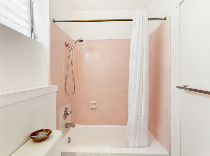 A bathroom with a pink-tiled shower, white curtain, showerhead, bathtub, and a small bowl on a toilet tank near a window.