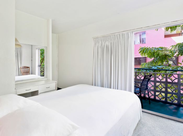 A bedroom with a white bed, a dressing table with a mirror, and a sliding door leading to a balcony with greenery outside.