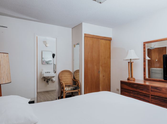 A neatly arranged hotel room with a bed, wooden furniture, a TV, a lamp, and a mirror. The bathroom is visible, with wicker chairs nearby.