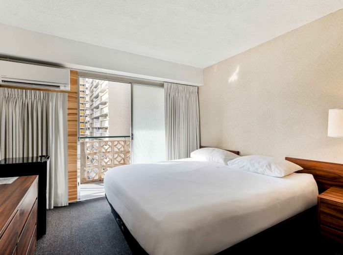 A neat hotel room features a large bed, wooden furniture, and a balcony with city views. The decor is minimal and modern.