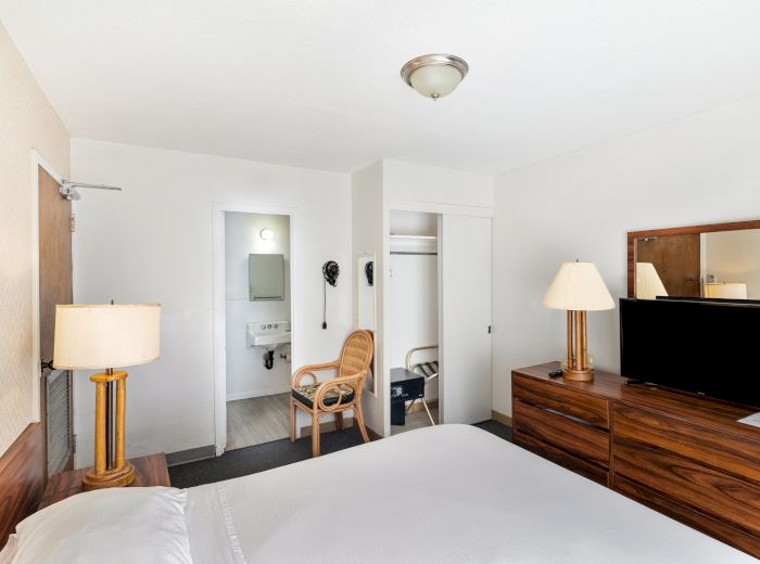 A hotel room with a bed, lamps, TV on a dresser, a mirror, and a chair near an open bathroom door in a minimalist setting.