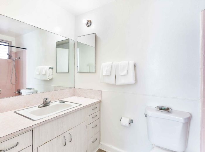 A clean bathroom features a sink, large mirror, towels, and a toilet with a small shelf, all in a white and light pink color scheme.