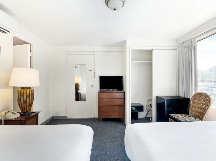 The image shows a hotel room with two beds, a TV, a lamp, a window, a closet, and a chair; the decor is simple and minimalist.