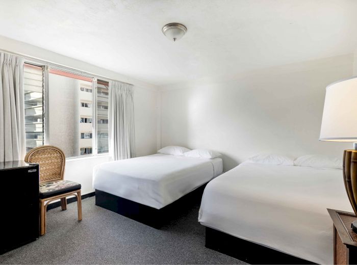 The image shows a hotel room with two double beds, a lamp, and a chair by a window, offering a simple and bright setting.