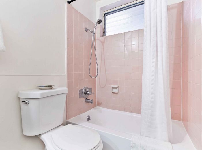 A bathroom with a white toilet, a bathtub with pink tiles, a showerhead, and a curtain is shown. A small window is above the shower.
