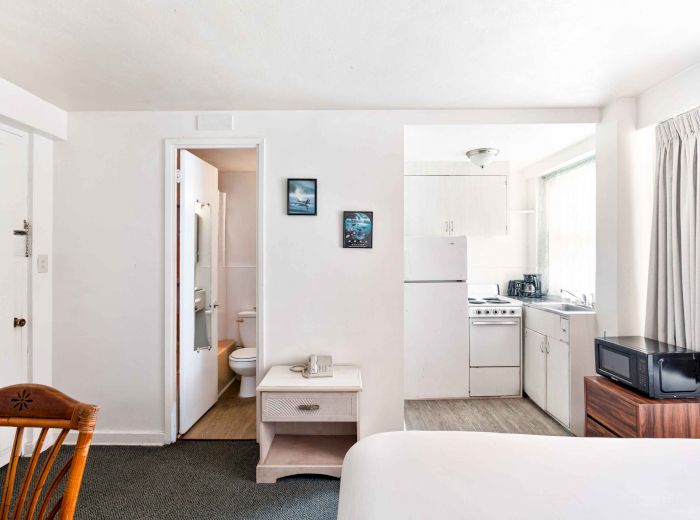 The image shows a compact room with a bed, small kitchen, bathroom, and wooden chair, creating a cozy, functional living space.