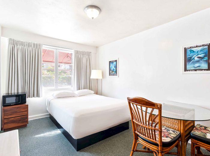 The image shows a bright hotel room with a bed, table, wicker chairs, a microwave, and two framed pictures on the walls.