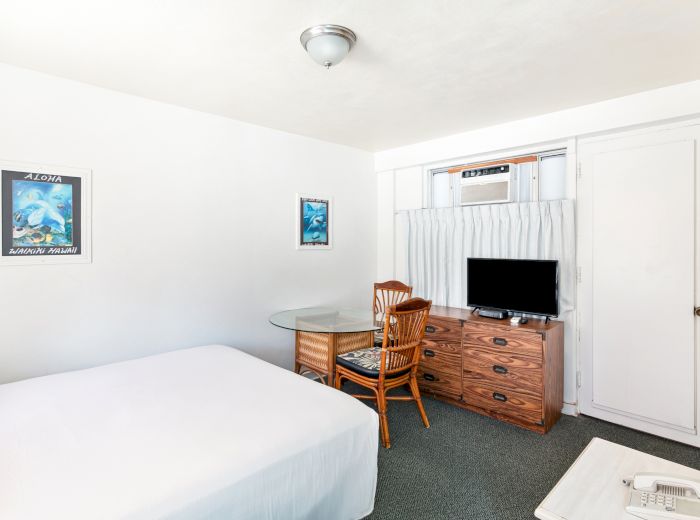 A small, simple room with a bed, a wooden chair, a dresser, a TV, and wall art, featuring minimalistic decor and light-colored walls.