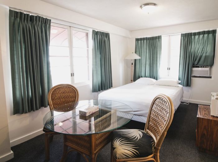 This image shows a cozy bedroom with a bed, green curtains, glass table with chairs, a wooden nightstand, and an air conditioner, all simply decorated.
