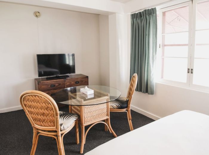 A room with a glass table, three chairs, a TV on a wooden dresser, and a bed, next to a window with green curtains.