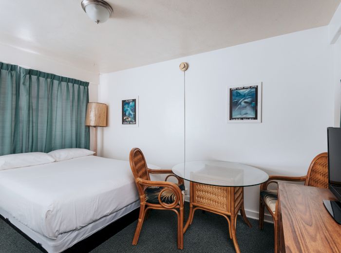 The image shows a hotel room with a bed, two wicker chairs, a glass-topped table, a TV on a wooden dresser, green curtains, and wall art.