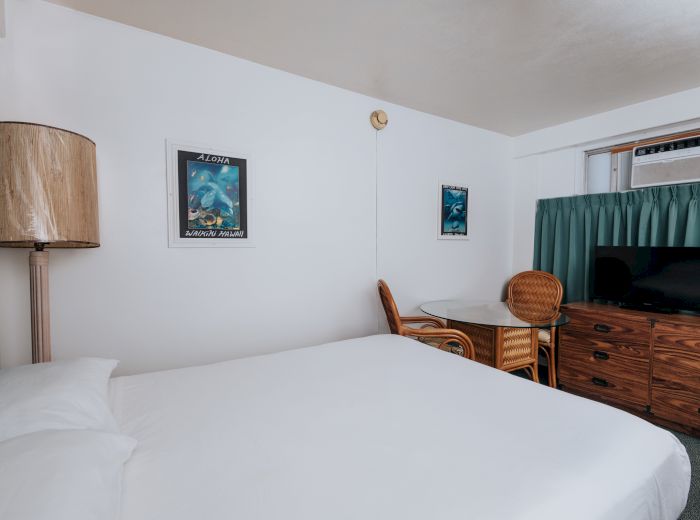 A hotel room with a bed, wicker furniture, framed pictures, a TV on a dresser, and an air conditioner, all in a cozy setting, ending the sentence.