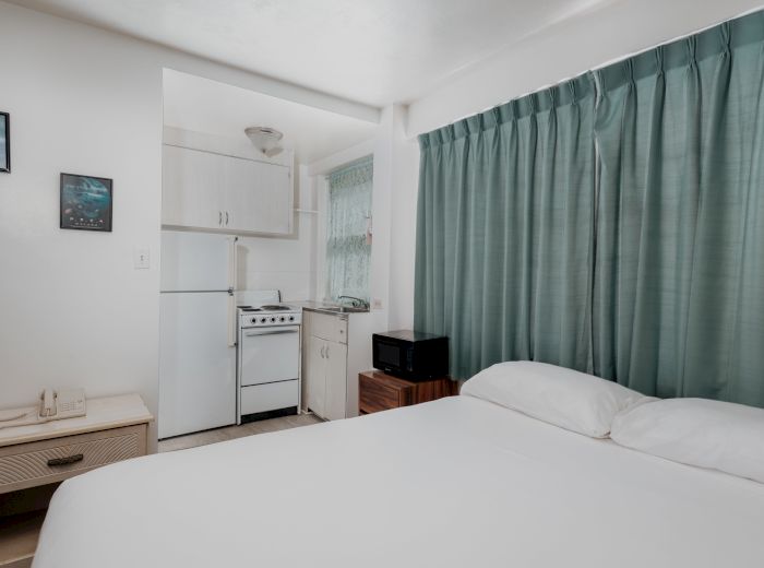 A small room features a bed, a kitchenette with fridge and stove, a microwave, green curtains, and two framed pictures on the wall, ending the sentence.