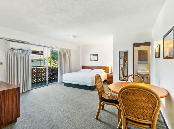 A bright hotel room features a bed, a small dining table with chairs, a TV stand, and a sliding glass door leading to a balcony.