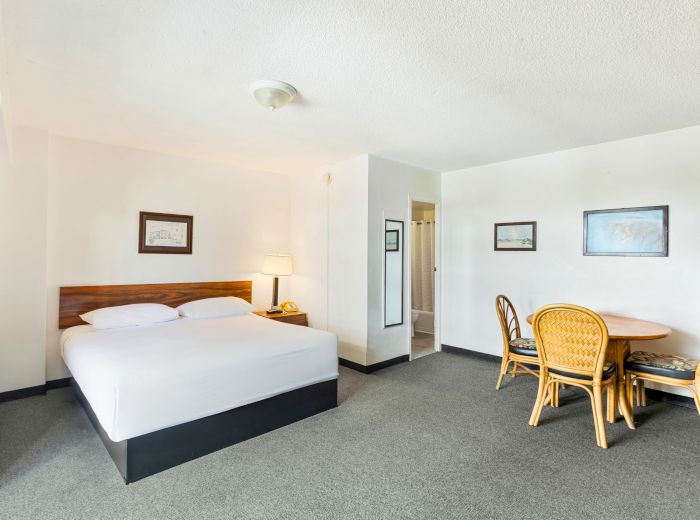 The image shows a hotel room with a bed, a small dining table with chairs, a lamp, and framed art on the walls.