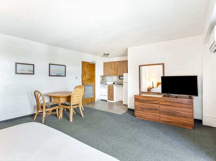 The image shows a simple room with a small kitchenette, dining table, TV, and bed in a minimalist style.