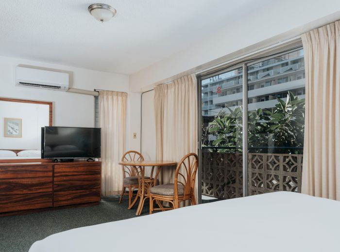 A hotel room with a bed, dresser with a TV, a small table with chairs, and a window with curtains leading to a balcony.