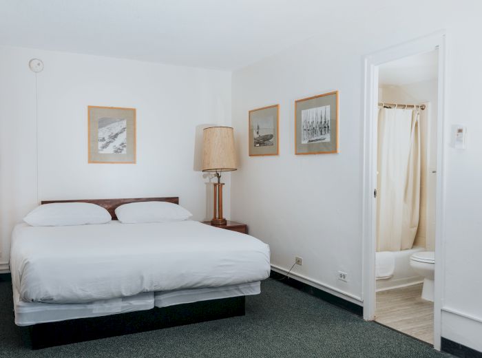 A small, minimalist bedroom with a double bed, bedside table and lamp, three framed pictures, and an open door leading to a bathroom.