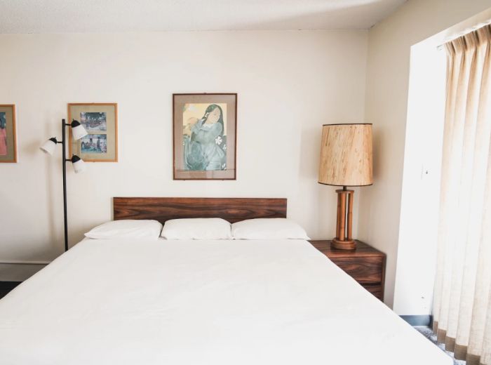 A bedroom with a large bed, three framed artworks on the wall, a floor lamp, a lamp on a nightstand, and a window with curtains ending the sentence.