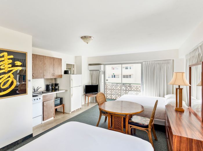 The image shows a hotel room with two beds, a kitchenette, a round table with chairs, a dresser with a lamp, and a wall decoration.