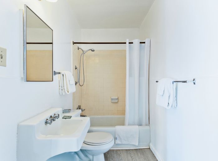 The image shows a clean bathroom with a sink, toilet, and shower curtain, alongside towels on racks and a wall mirror.