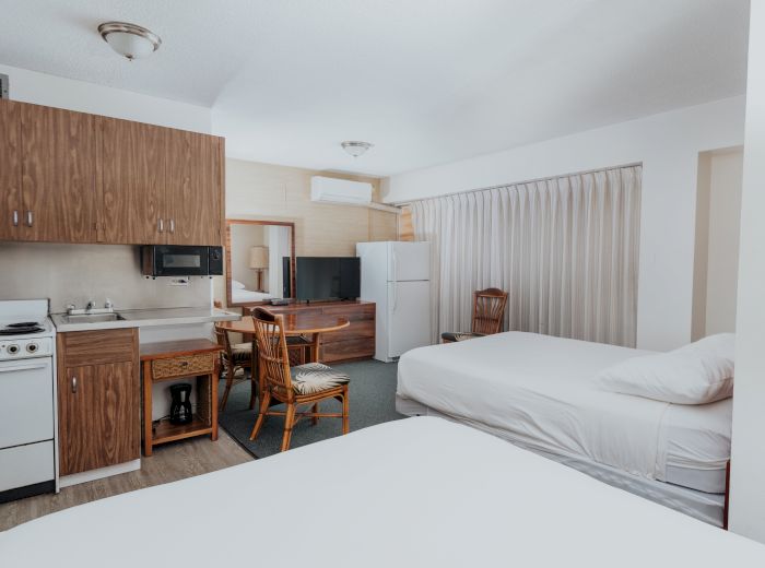 This image shows a room with two beds, a small kitchen area, a mini fridge, a microwave, and a table with two chairs, featuring wooden decor.