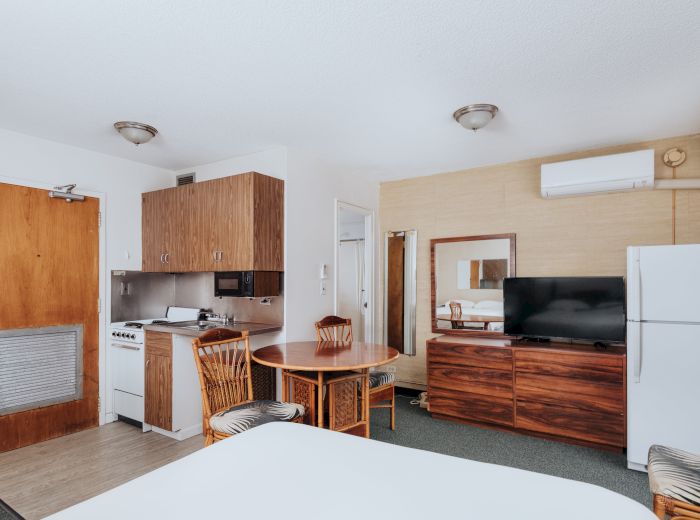 A hotel room with a bed, a kitchenette, a dining table with chairs, a TV, a refrigerator, and a door. The decor is simple and functional.