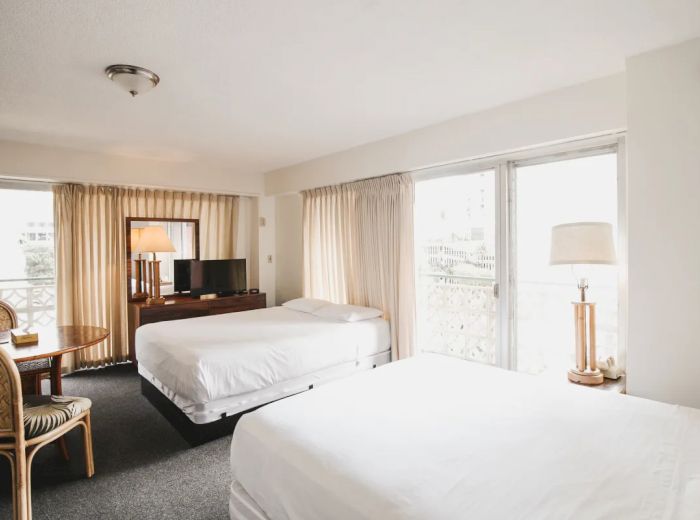 The image shows a bright hotel room with two beds, a TV, a lamp, a small table with chairs, and large windows with curtains.