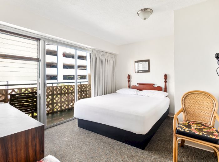 A simple bedroom with a double bed, balcony view, wooden dresser, rattan chair, and wall-mounted retro telephone.