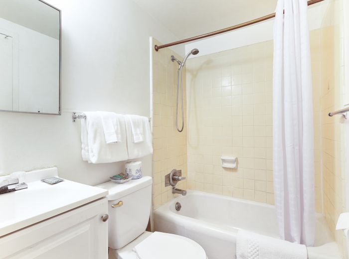 A clean bathroom with a bathtub, white shower curtain, towels, and mirror. The tiles are beige, and there's a toilet with amenities.