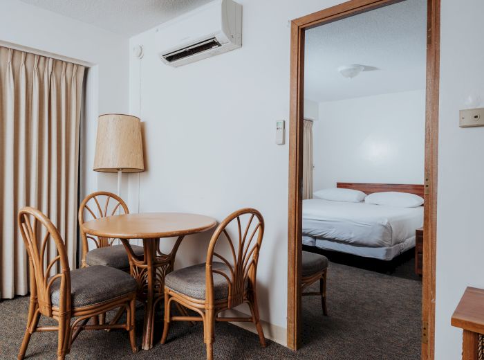 A small dining area with a round table and four chairs is next to an open door leading to a bedroom with a double bed and minimalist decor.