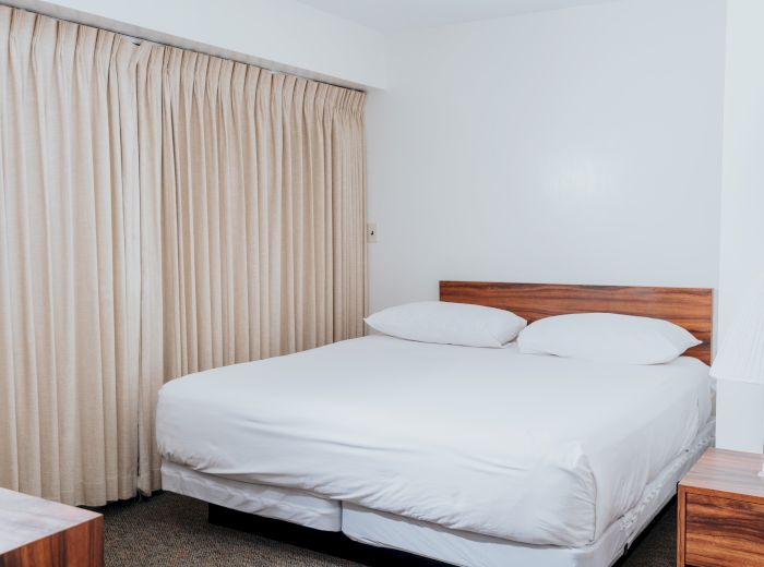 The image shows a minimalist bedroom with a large bed, white linens, beige curtains, and a wooden bedside table with a lamp.