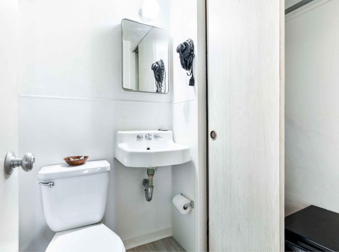 A small bathroom with a toilet, sink, mirror, wall-mounted hairdryer, and a wooden door leading to a closet is shown.