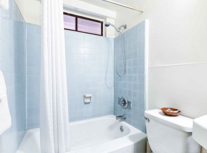 The image shows a bathroom with a white bathtub, blue tiled walls, showerhead, white curtain, and a toilet with items on the tank lid.