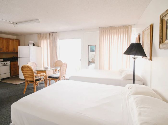 A hotel room with two beds, a kitchenette, a table with chairs, a refrigerator, and a lamp, adorned with paintings on the walls.