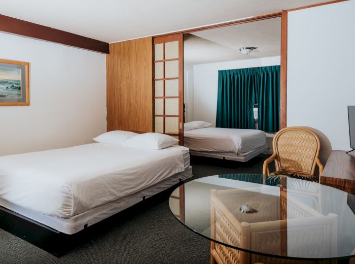 A hotel room with two beds, a glass table, a TV, a wooden chair, blue curtains, and a framed picture on the wall.