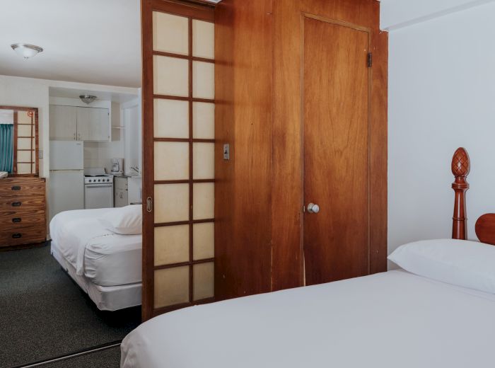 The image shows a bedroom with a wooden sliding door separating it from another room with a dresser, bed, and a small kitchen area in the background.