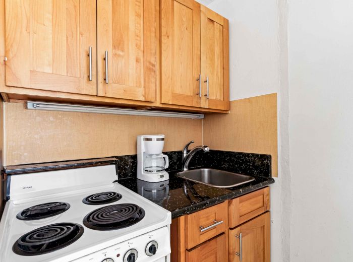 The image shows a compact kitchen with wooden cabinets, an electric stove, a coffee maker, and a sink next to a granite countertop.