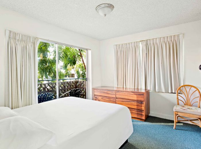 A bright bedroom with a bed, chest of drawers, chair, and windows covered with curtains that overlook greenery outside.