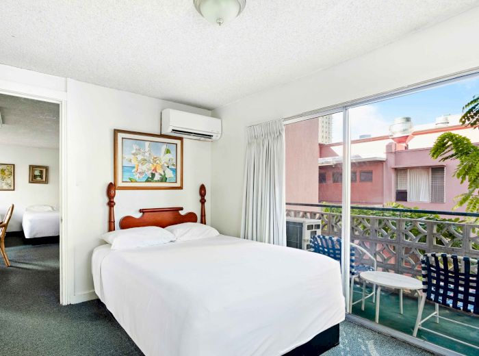 The image shows a bright room with a bed, painting, air conditioner, and sliding glass doors leading to a balcony with chairs.