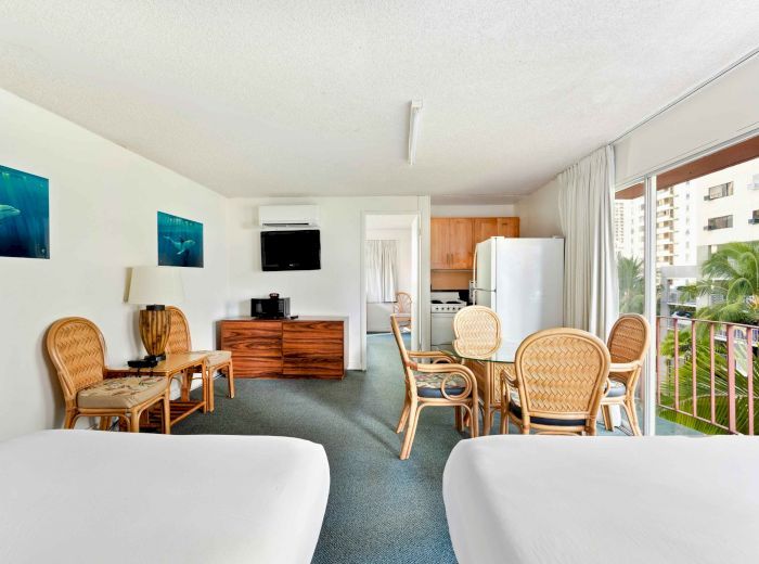 The image shows a bright hotel room with two beds, a seating area, a dining table, a small kitchen, and a balcony with a view.
