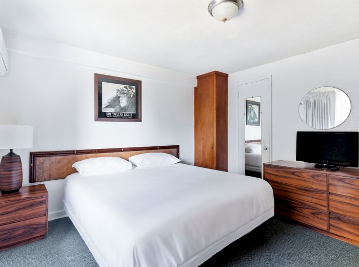 A neatly arranged bedroom with a bed, a wooden dresser, a TV, a mirror, a lamp, and wall art; all in a minimalist style.