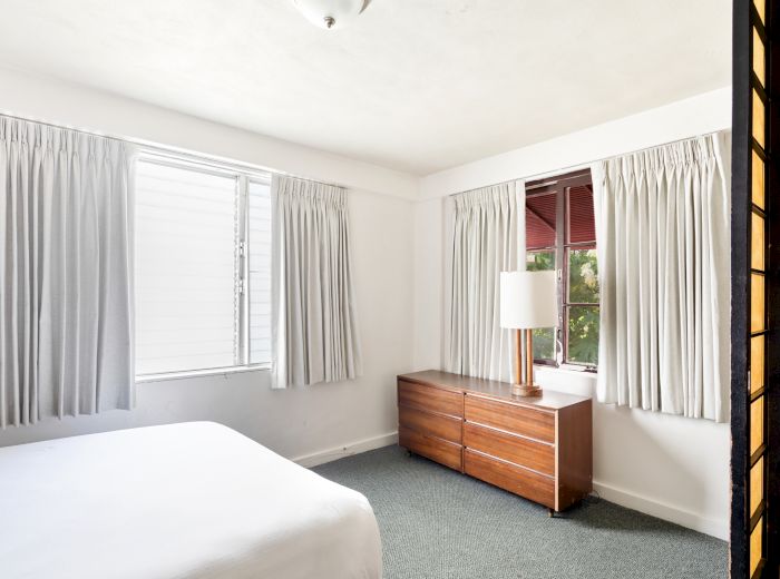 A minimalist bedroom with a bed, dresser, lamp, and curtains covering windows; the room is bright and simple in design.