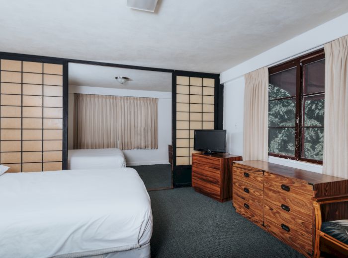 The image shows a bedroom with a bed, a wooden dresser with a TV, and a separate area with another bed, separated by sliding doors.