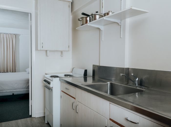 A small kitchen with a sink, stove, and upper shelves holding pots and pans, with a doorway leading to a bedroom with a bed.
