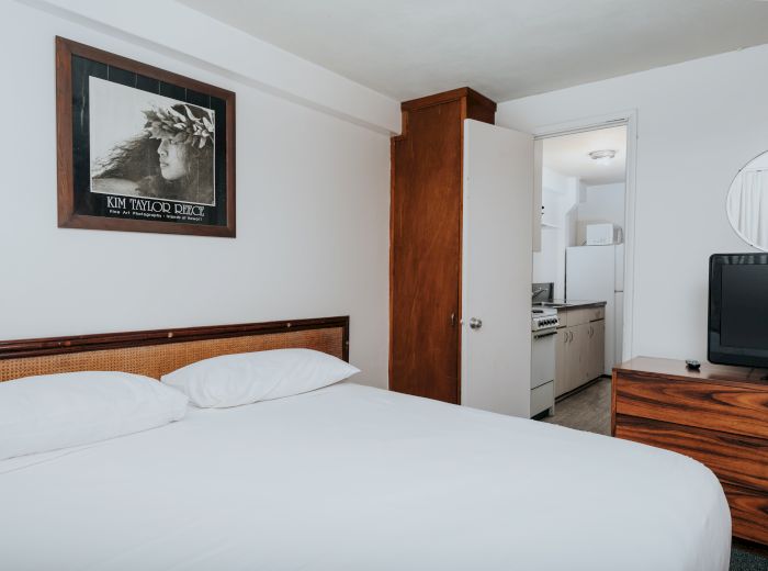 A minimalist bedroom with a bed, a framed picture, a wooden dresser with a TV, and an open door leading to a small kitchen area.